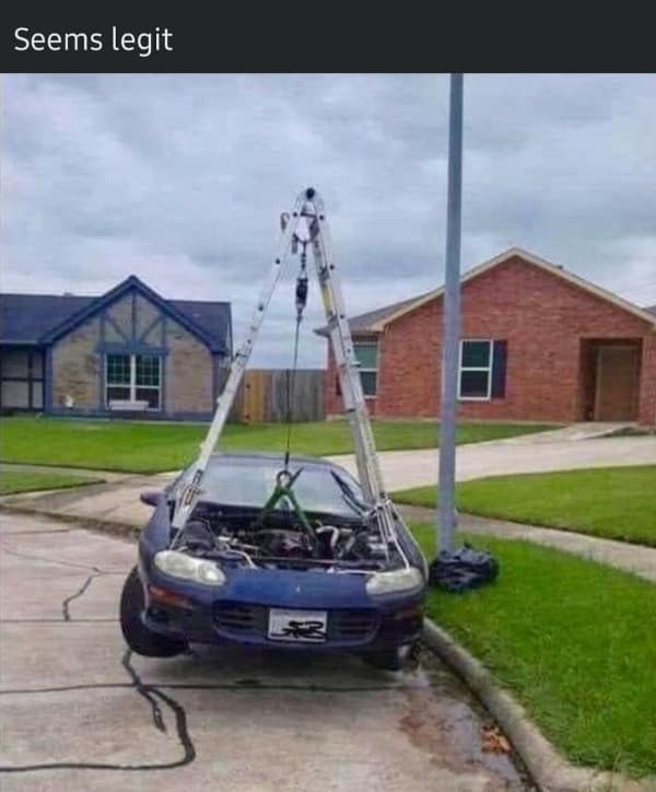A hilarious "redneck engineering" funny fail showing a blue car parked on a street curb where someone is dangerously attempting to lift the heavy engine out using nothing but a standard aluminum step ladder and a pulley.