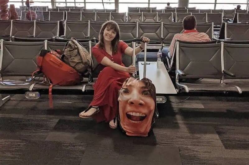 Woman sitting in an airport lounge next to a suitcase printed with a giant screaming face.
