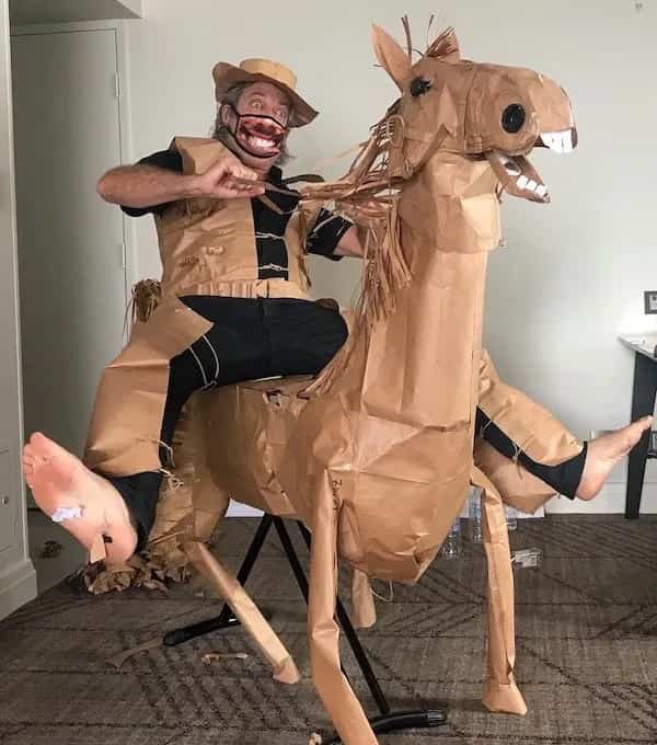 A man in a brown paper horse costume with a realistic smiling mouth mask posing indoors.