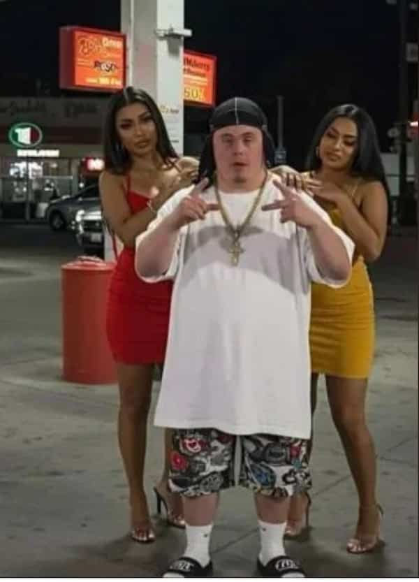 A man with Down syndrome posing confidently at a gas station between two glamorous women.