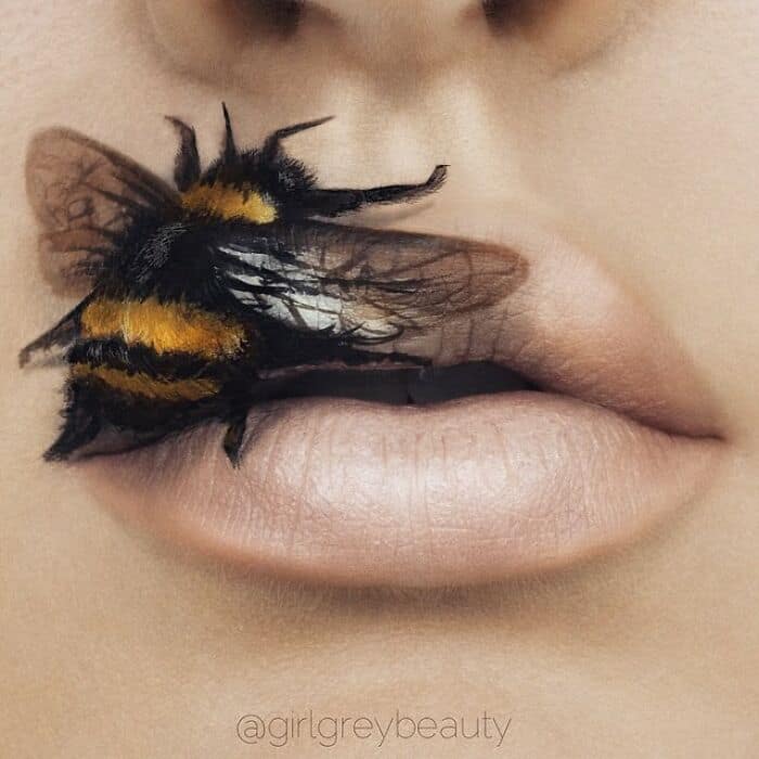 Mind-bendingly realistic and amazing lip art featuring a 3D-painted, fuzzy bumblebee with translucent wings resting gently on the corner of soft, natural lips, a testament to the surreal talent behind Andrea Reed lip art.