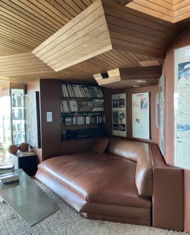 A vintage 70s-style masculine home decor reading nook featuring a massive built-in brown leather lounge bed, wood-paneled angular ceilings, and packed bookshelves.