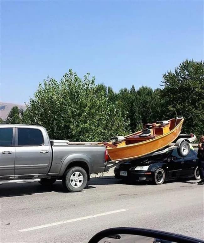A disastrous driving fail captured in a funny meme showing a large wooden boat on a trailer completely crushing the roof of a black sedan behind a pickup truck, a perfect chaotic visual for a meme dump.