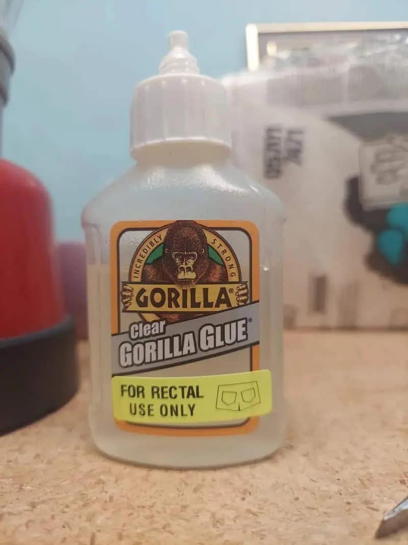 An absolutely chaotic funny meme showing a bottle of incredibly strong clear Gorilla Glue that someone has maliciously pranked by slapping a bright yellow pharmacy warning sticker on it that reads "FOR RECTAL USE ONLY," making it a perfect cursed addition to a meme dump.