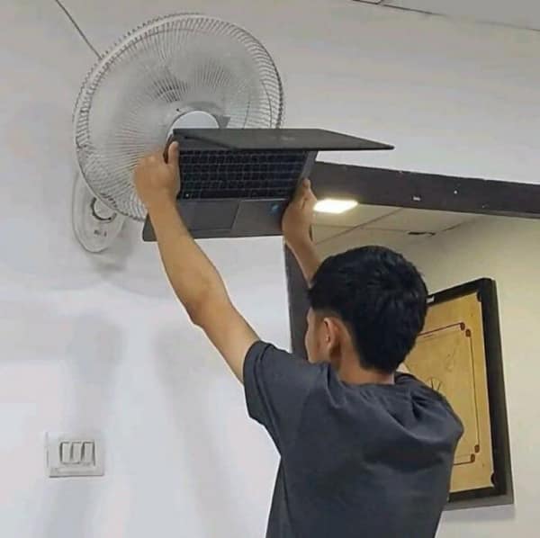 A nerd meme showing a gamer holding a laptop up to a wall-mounted fan to prevent overheating.