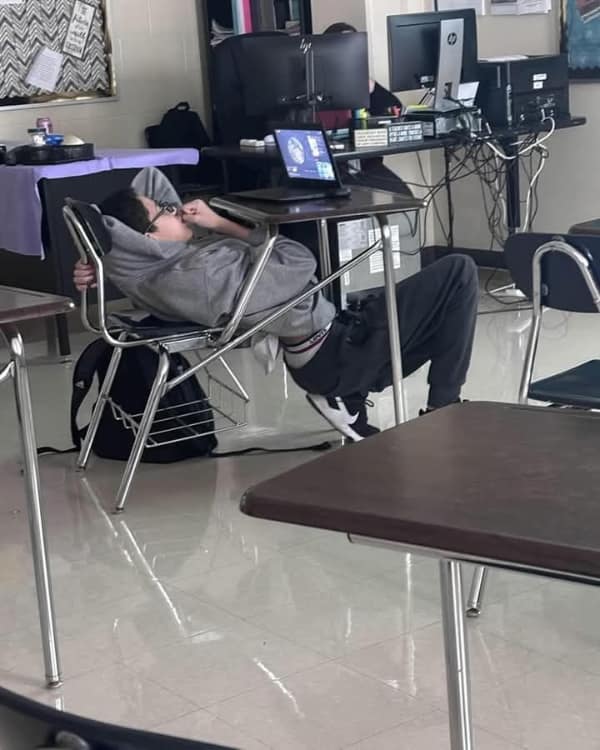 A nerd meme photo of a student sleeping comfortably while suspended contorted under a school desk.