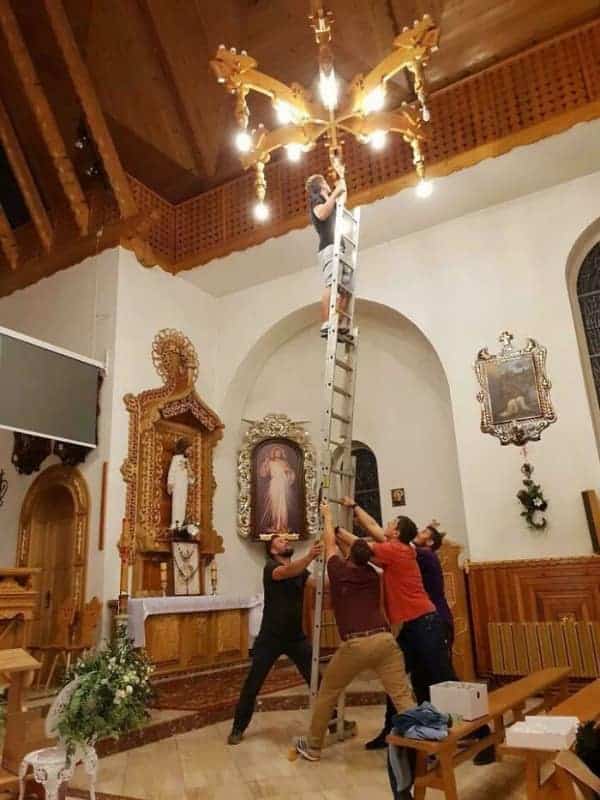 A community effort osha meme showing five men holding a massive extension ladder vertically in a church so one brave soul can change a chandelier bulb.
