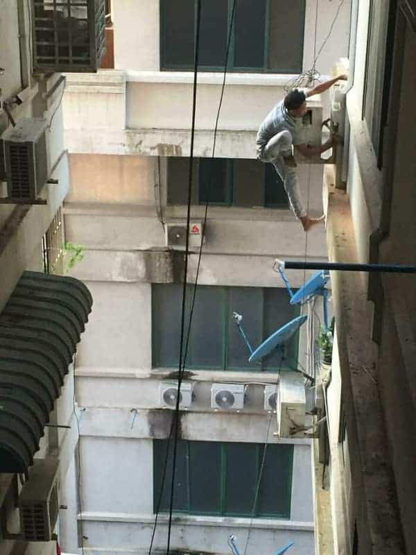 A terrifying osha meme capturing a barefoot technician balancing precariously on a high-rise ledge to install an AC unit with zero fall protection.