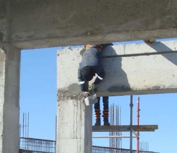 An anxiety-inducing osha meme of a construction worker clinging to a concrete beam like a koala to finish a job high above the ground.