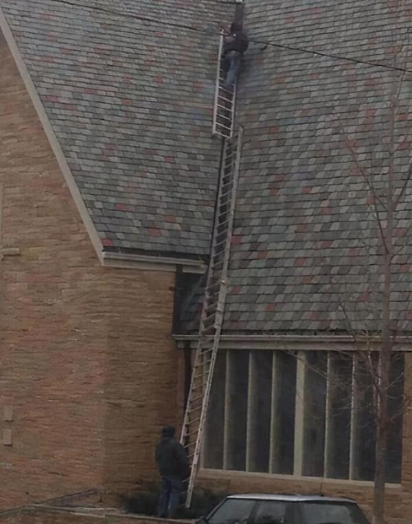 A dizzying osha meme of a roofer climbing an impossibly long, steep ladder setup that extends all the way up a high-pitched roof without visible safety gear.