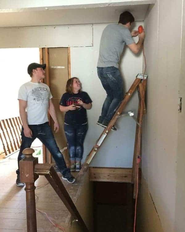 A funny osha fail depicting two guys improvising a ladder setup on a staircase by balancing one leg on the banister while a woman watches in apparent concern.
