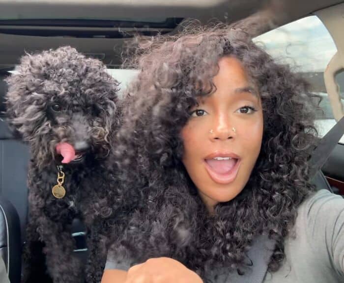 A woman with voluminous dark curly hair smiling next to her black poodle with identical curls.