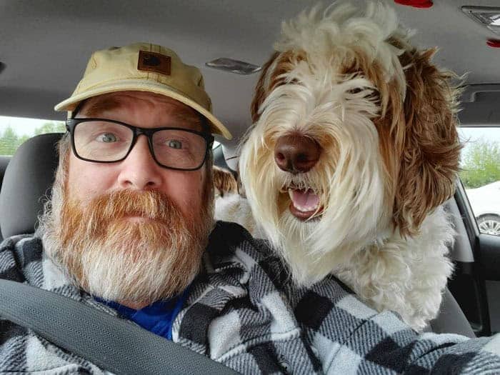Bearded man in a flannel shirt and cap next to a scruffy white and tan dog.