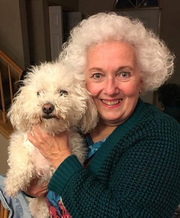 Older woman with white curly hair smiling next to a white curly-haired bichon frise dog.