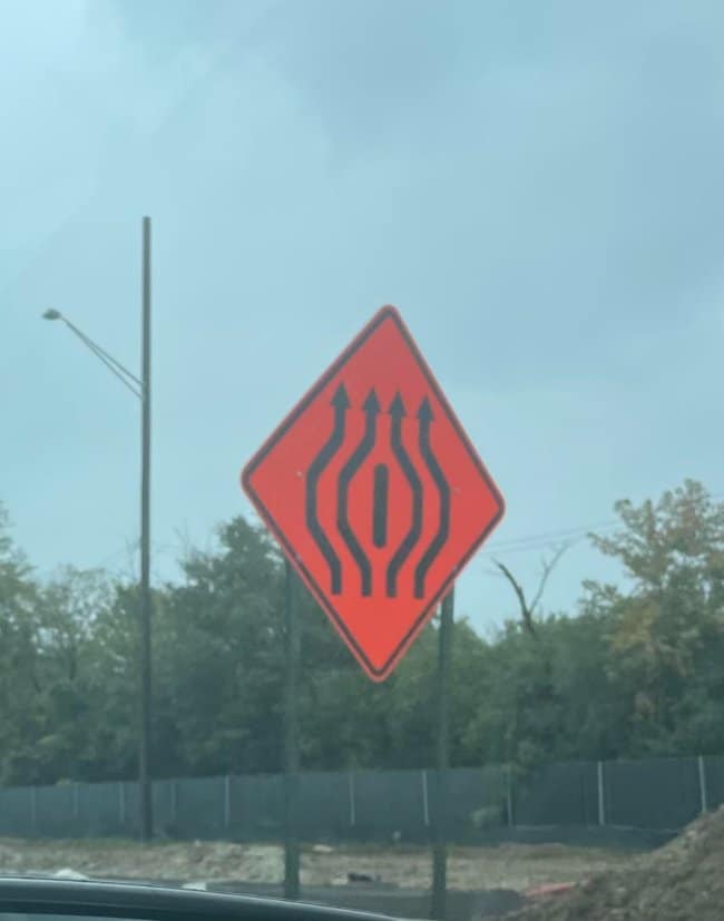 A traffic warning sign on a road featuring five arrows merging around a center island, resulting in a highly accidentally nsfw shape and a classic unfortunate design fail.