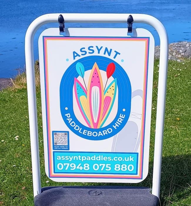 An advertisement sign for "Assynt Paddleboard Hire" showcasing an unfortunate design where overlapping colorful paddleboards and paddles create an accidentally nsfw silhouette.