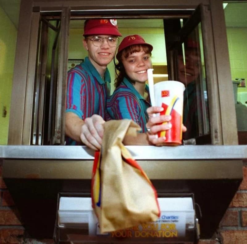 A vintage McDonalds image of two employees smiling in the drive-thru window wearing teal and red striped 90s uniforms.