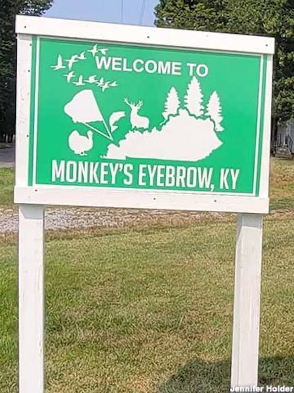 Green Welcome to Monkey's Eyebrow Kentucky sign featuring a white map outline and wildlife icons.