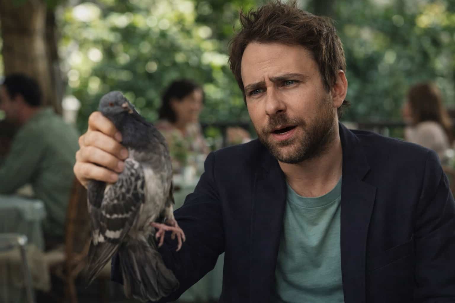 Charlie Day lookalike holding a pigeon while looking confused in an outdoor restaurant setting.