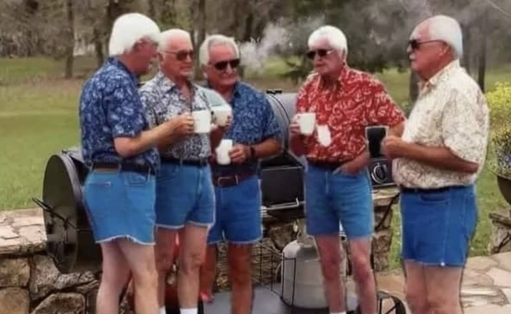 Five elderly men wearing matching floral shirts and denim cut-off shorts standing by a grill.