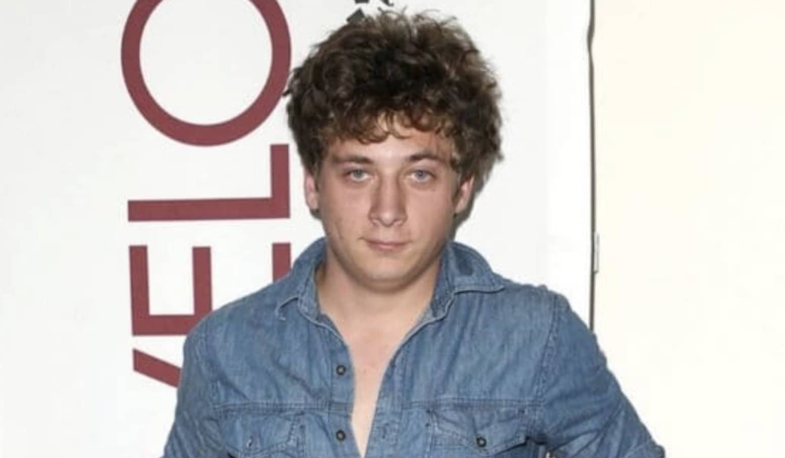 Young Jeremy Allen White with curly hair wearing a denim shirt at a press event.