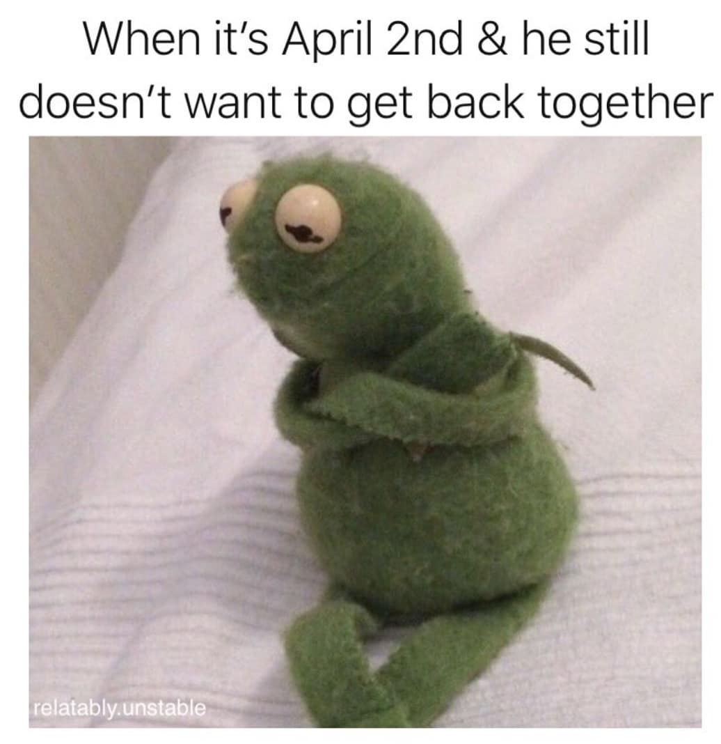 A petty April Fools Day meme showing a small, grumpy green felt puppet sitting with its arms crossed in bed. The caption reads: "When it's April 2nd & he still doesn't want to get back together," poking fun at people who use the holiday to test a partner's feelings and then try to take it back.