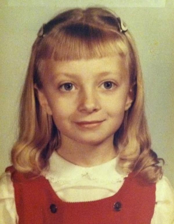A young girl in a school portrait with a layered 80s haircut. She features micro-bangs and hair pinned back at the temples with clips, transitioning into longer, feathered waves that hit her shoulders.