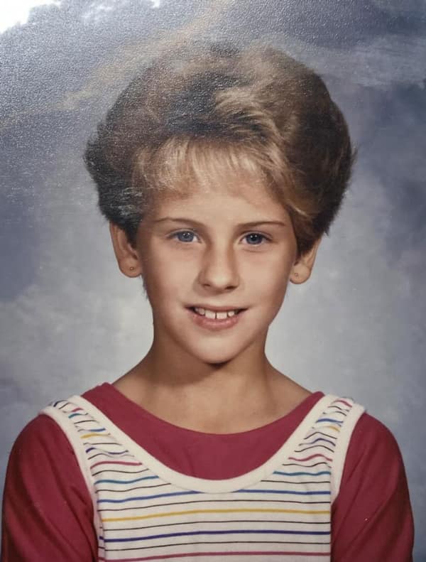 The ultimate "mall hair" 80s haircut on a young girl. This style features extreme volume on top, achieved through heavy teasing, with feathered layers swept back and thin, wispy bangs across the forehead.