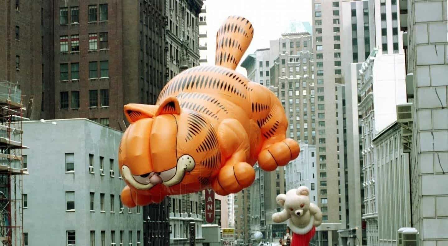 A vintage nostalgia meme photo from a 90s Macy's Thanksgiving Day Parade, featuring a massive, grinning Garfield the Cat balloon floating between New York City skyscrapers.