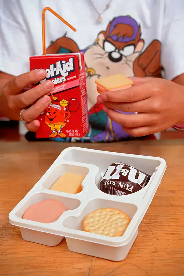 classic lunchroom staple in these 90s pictures showing a person putting together an iconic Lunchables meal, featuring crackers, cheese, ham, a fun-size M&M's pack, and a cherry Kool-Aid Jammers juice box.