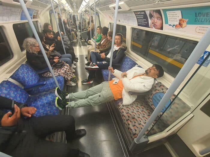 A man sprawls out for a nap across an entire row of seats on a crowded London Underground train, illustrating the complete lack of spatial awareness from annoying people on public transit.