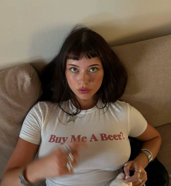 Young woman wearing a tight white t-shirt that simply reads "Buy Me A Beer?" in red text.