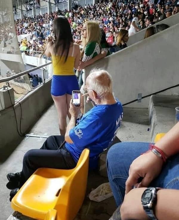 Older man at a stadium holding his phone up to take a blatant photo of a woman's backside.