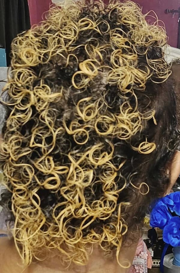 A heavily damaged bad haircut featuring a close-up of dark curly hair severely fried by bleach, resulting in stiff, bright yellow ringlets that look exactly like scattered, dry rubber bands.