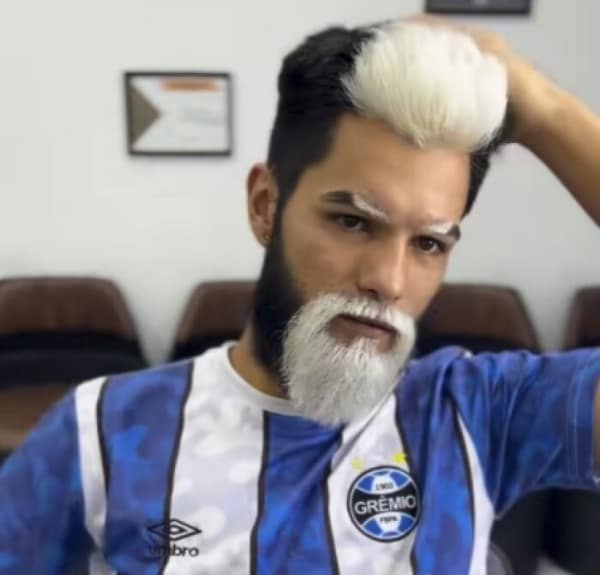 A highly stylized and bizarre bad haircut where a man has dyed the top swoop of his hair, his eyebrows, and his sharply lined goatee a bright platinum white, contrasting harshly with his dark brown hair.