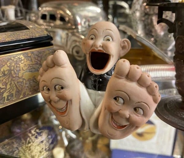 A peak funny thrift store find: a cursed ceramic figurine featuring a bald man with a wide-open, screaming mouth flanked by two large upright feet that have detailed, smiling faces painted onto the soles.