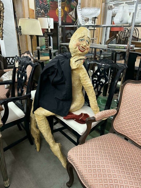 An unsettling life-sized sculpture of a lumpy, yellow-skinned figure with a manic grin and painted-on eyes, slumped in a white chair and draped in a black jacket amidst a furniture section.