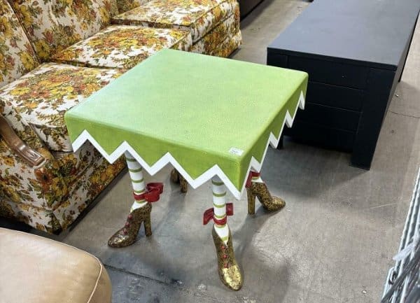 small square end table with a lime green top and zig-zag edges, supported by four legs wearing striped stockings and gold glitter high-heeled boots, sitting in a furniture aisle.