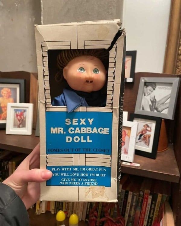 A knock-off Cabbage Patch-style doll in a battered box labeled "Sexy Mr. Cabbage Doll: Comes Out of the Closet," with a tagline encouraging the buyer to "Give me to anyone who needs a friend."