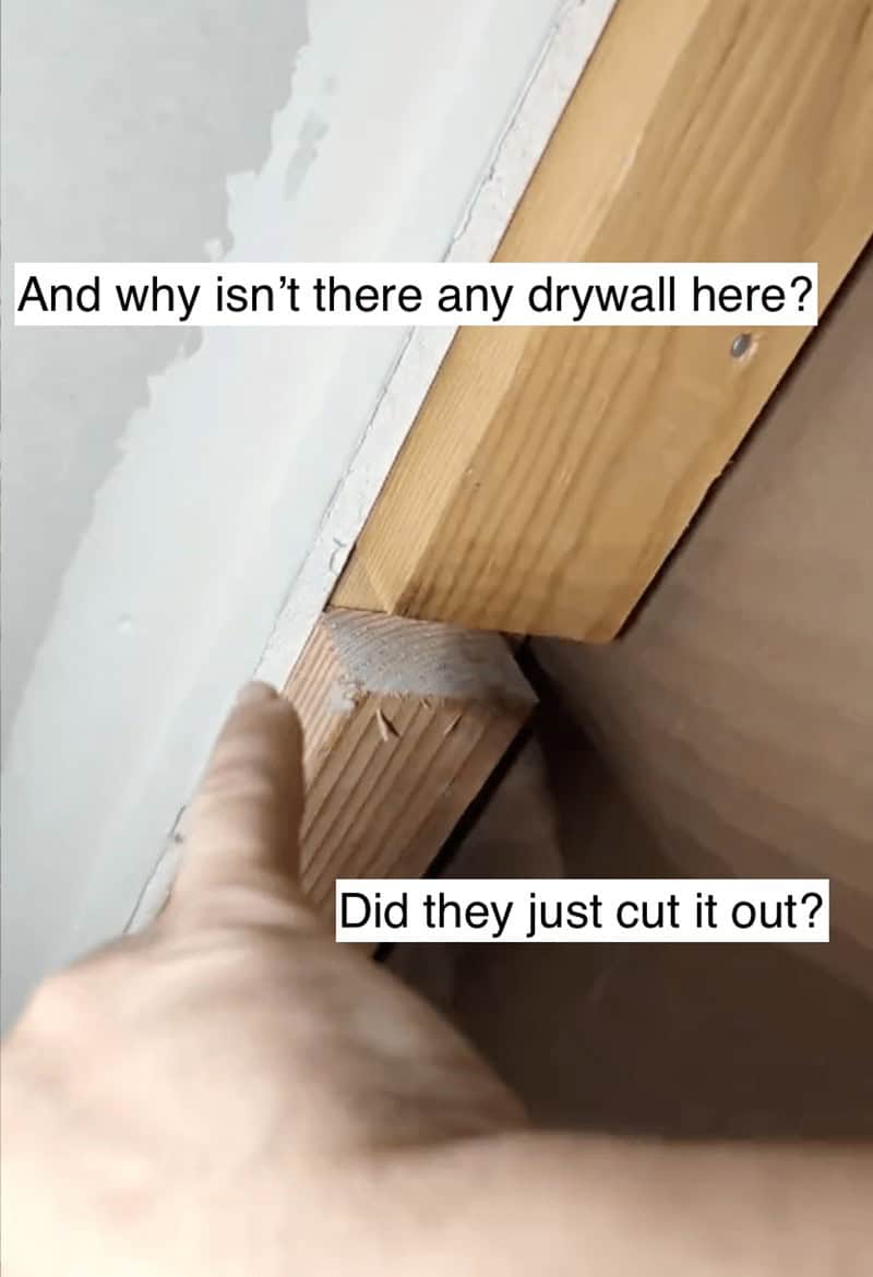 Hand pointing at a gap where drywall was cut away exposing the wooden wall studs.