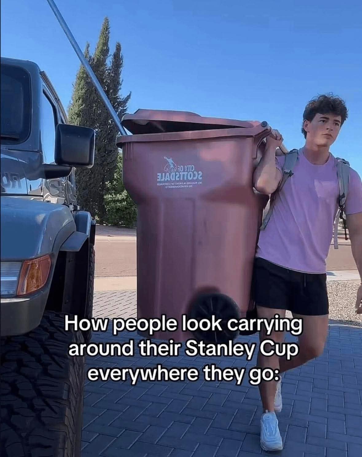 classic meme parodying the Stanley Cup tumbler obsession, showing a man walking down a suburban street carrying a literal full-sized city trash bin on his shoulder to mock the massive size of the viral water bottles.