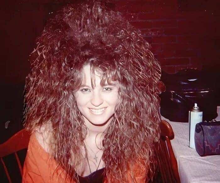 Woman with dark reddish-brown hair featuring a vertical, gravity-defying teased crown and long curls.