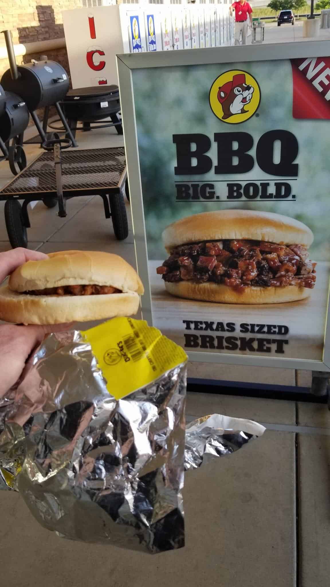 A hand holding a small, flat Buc-ee's brisket sandwich next to a large, overflowing advertisement.