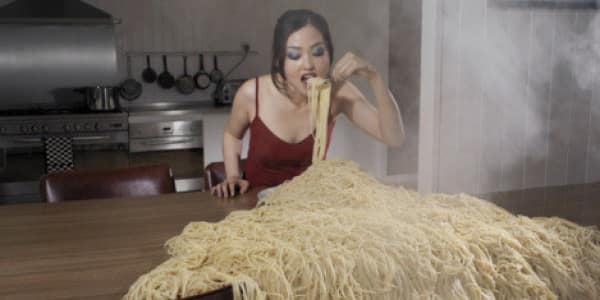 Woman aggressively eating an absolutely towering mountain of steaming spaghetti directly off a kitchen table. A wildly relatable food meme for pasta lovers