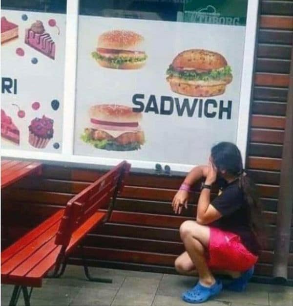 Man dramatically crouching and crying outside a fast food window next to a typo on a menu sign that accidentally advertises a Sadwich.