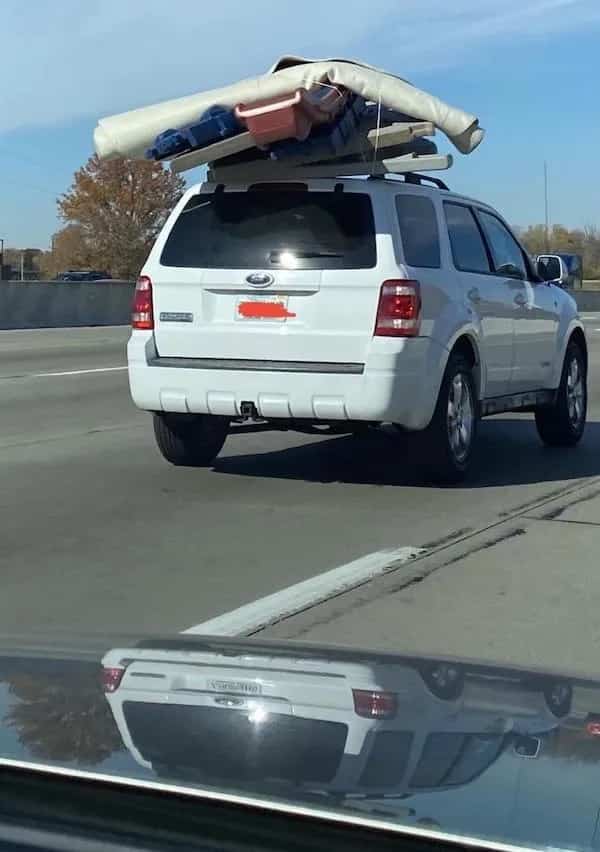 White SUV driving on a highway with an incredibly precarious and oversized load of furniture on its roof.