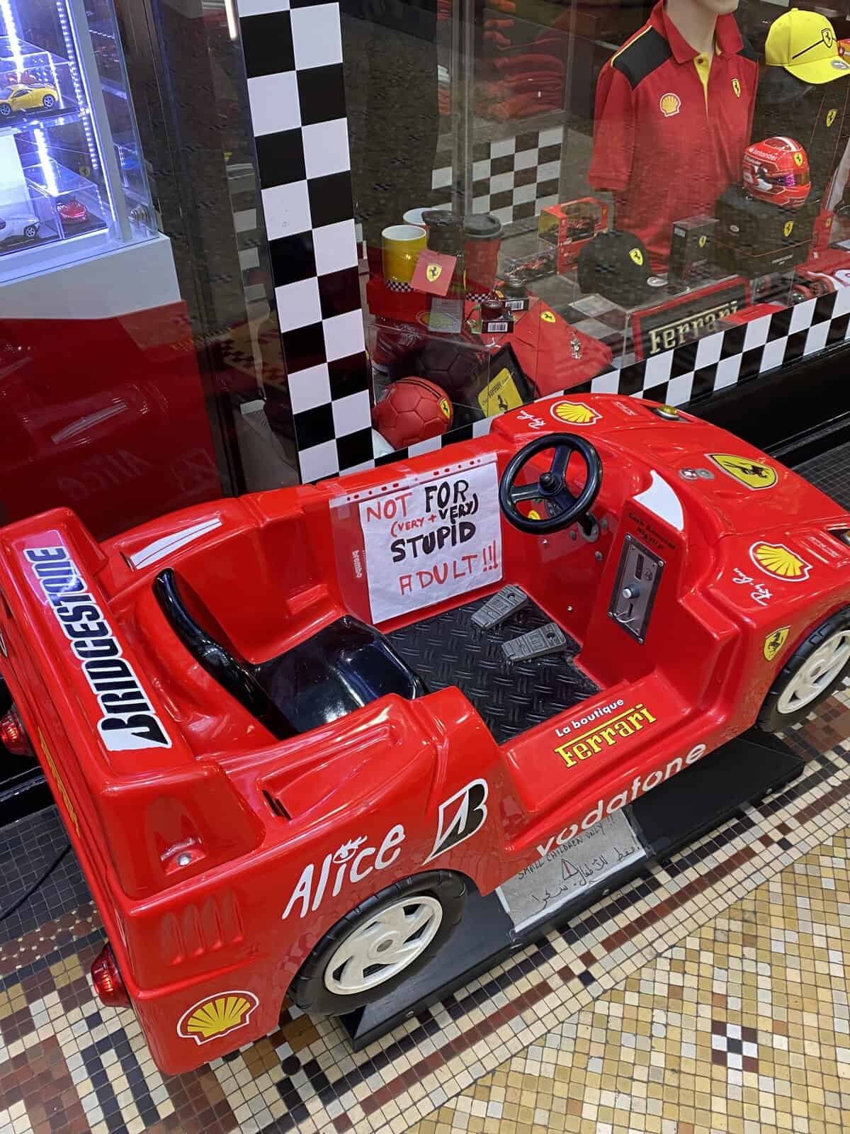 miniature red Ferrari kiddy car warns against misuse with a taped-on sign stating it is "NOT FOR (VERY+VERY) STUPID ADULT!!!".