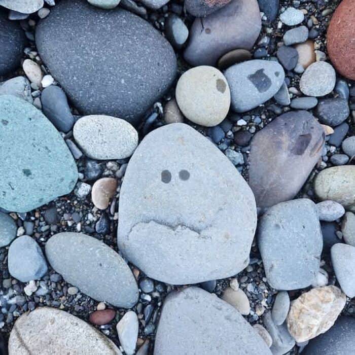 inanimate objects with faces showing a grey rock on a pebble beach, where two dark spots and a jagged crack create a perfectly unimpressed, frowning expression.