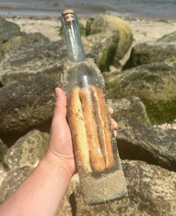Hand holding a glass message in a bottle found at the beach mysteriously containing two fully assembled hot dogs. Absurd and unexplained funny memes
