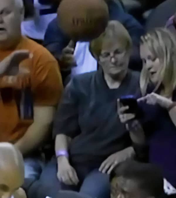 A basketball mid-flight about to hit a woman looking at her phone in the stands.